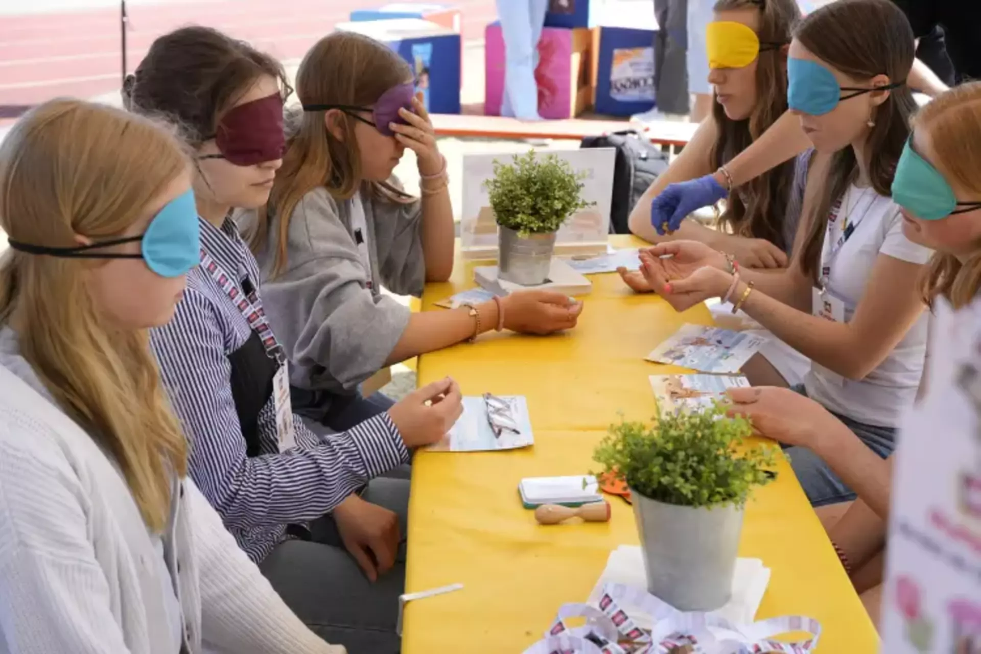 Auf dem Bild sieht man eine Gruppe junger Menschen, die an einem gelben Tisch sitzen – alle mit bunten Augenbinden. Es handelt sich um eine Station der Schwartau-Olympiade, vermutlich den „Schmeckathlon“, bei dem eine Blindverkostung stattfindet. Eine Person mit blauer Einweghandschuhen reicht gerade ein Produkt zum Probieren. Vor den Teilnehmenden liegen kleine Flyer, vermutlich zur Orientierung oder für Notizen. Die Szene zeigt konzentrierte Gesichter und eine ruhige, neugierige Atmosphäre. Durch die Augenbinden stehen Geschmack und Sinne im Mittelpunkt – ein kreativer Ansatz, um Barrieren spielerisch zu thematisieren und Erlebnisse zu schaffen, die verbinden. Im Vordergrund liegen wieder Schwartau-Lanyards, außerdem Topfpflanzen als freundliche Deko – alles liebevoll gestaltet. Das Bild strahlt Gemeinschaft, Teilhabe und Inklusion aus.