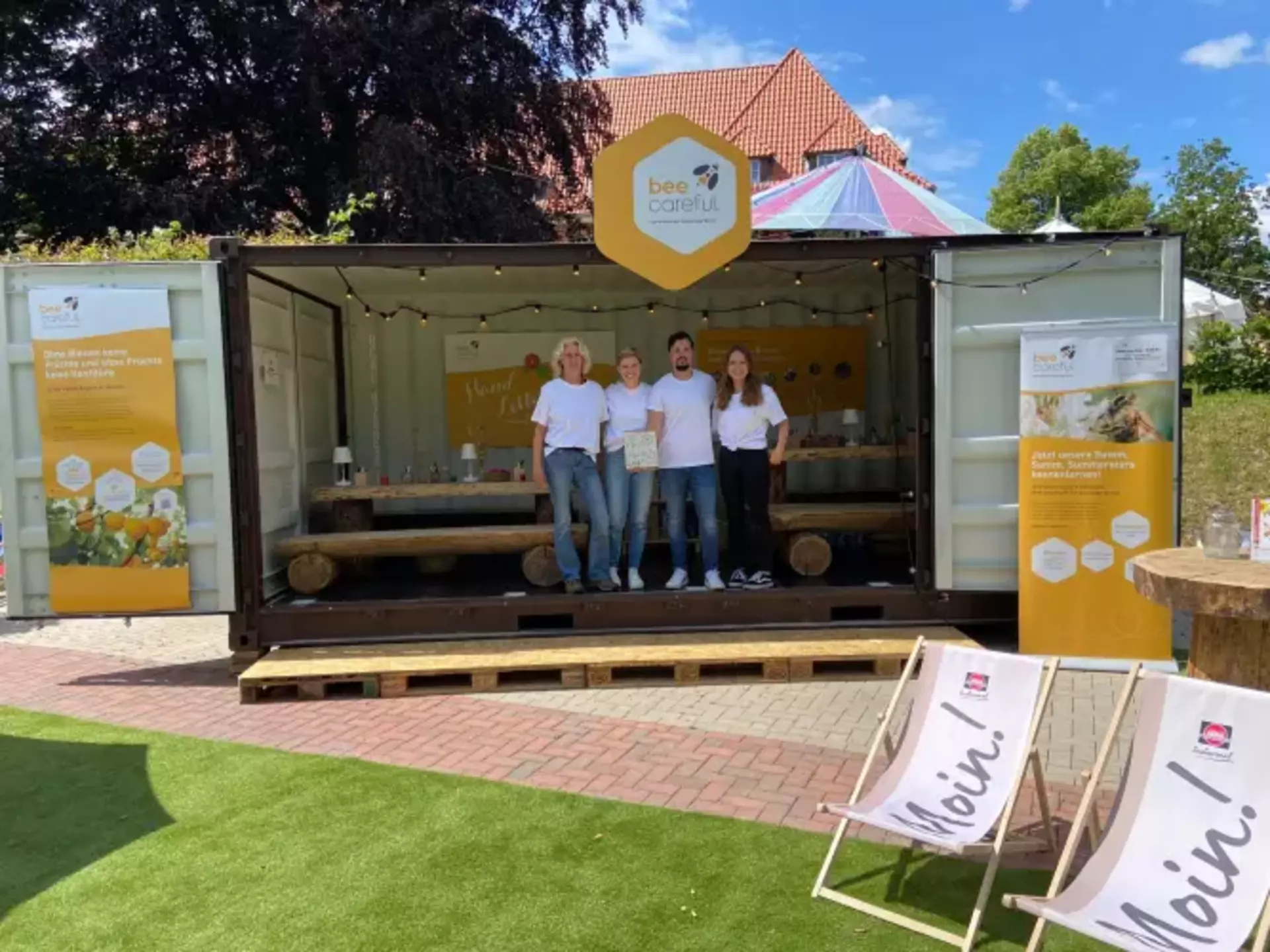 Man sieht drei Frauen und einen Mann gemeinsam vor einem großen geöffneten Stand stehen. An den Seiten sind große Infotafeln über das Thema Bienengesundheit. In der Mitte des Standes hängt ein großes Schild mit dem bee careful Logo. Vorn stehen Liegestühle mit dem Aufdruck Moin und dem Schwartau Logo. Es ist blauer Himmel zu sehen.