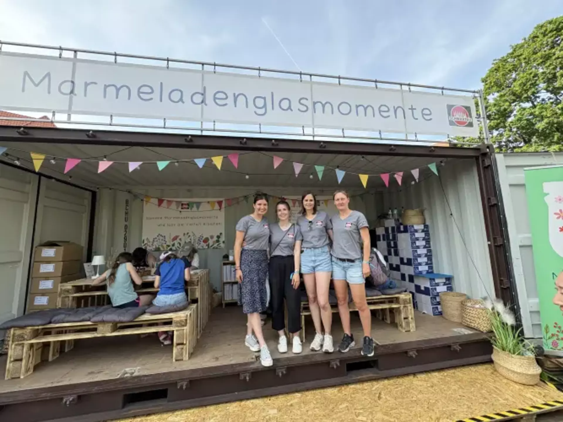 Man sieht vier junge Frauen in einheitlichen T-Shirts mit bunten Marmeladengläsern und Schwartau Logo, die am Eingang eines geöffneten Schiffscontainers stehen. Über ihnen sieht man ein großes Banner mit der Aufschrift Marmeladenglasmomente und eine bunte Girlande. Im Hintergrund sitzen Kinder an Holzmöbeln und malen.