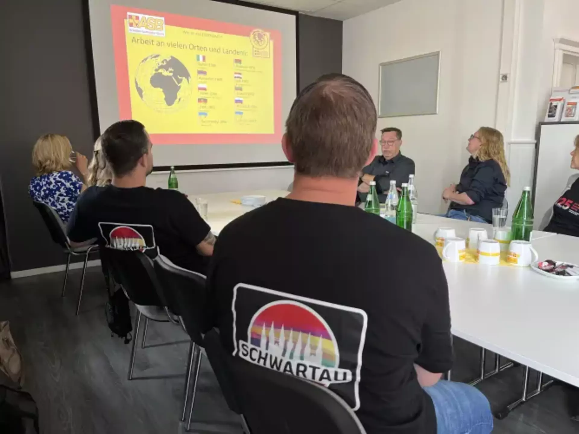 Das Foto zeigt sowohl Schwartau Kolleg*innen, als auch ASB Mitarbeiter*innen an einem Tisch. Alle Personen schauen auf eine Leinwand, an welcher eine Präsentation über die ASB gezeigt wird.