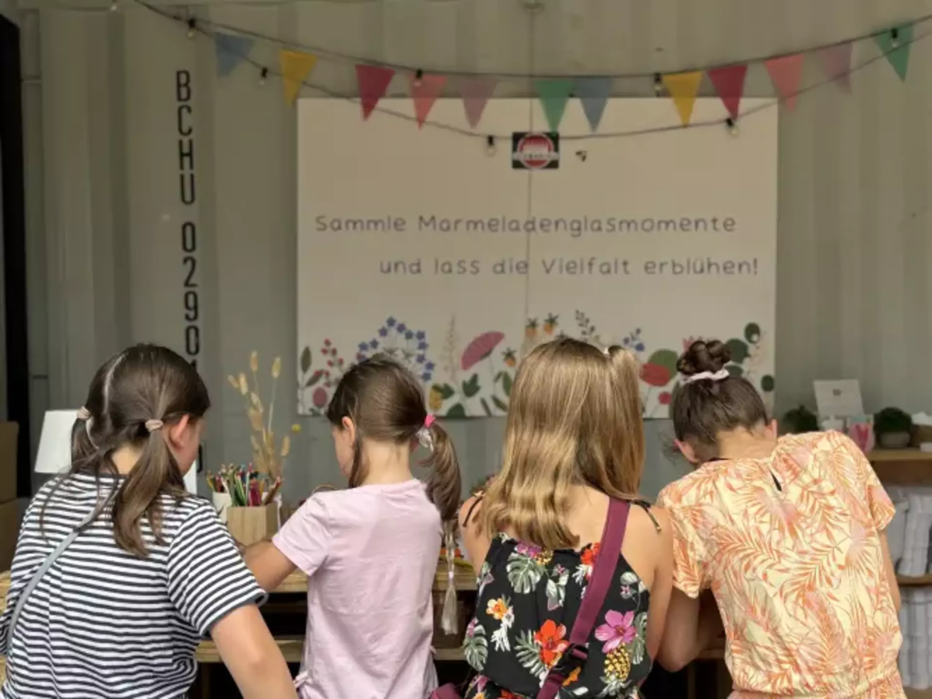 Man sieht vier Mädchen von hinten fotografiert an einem Holztisch sitzen und malen. Im Hintergrund hängt eine bunte Girlande und ein Schild mit der Aufschrift "Sammle Marmeladenglasmomente und lass die Vielfalt erblühen"
