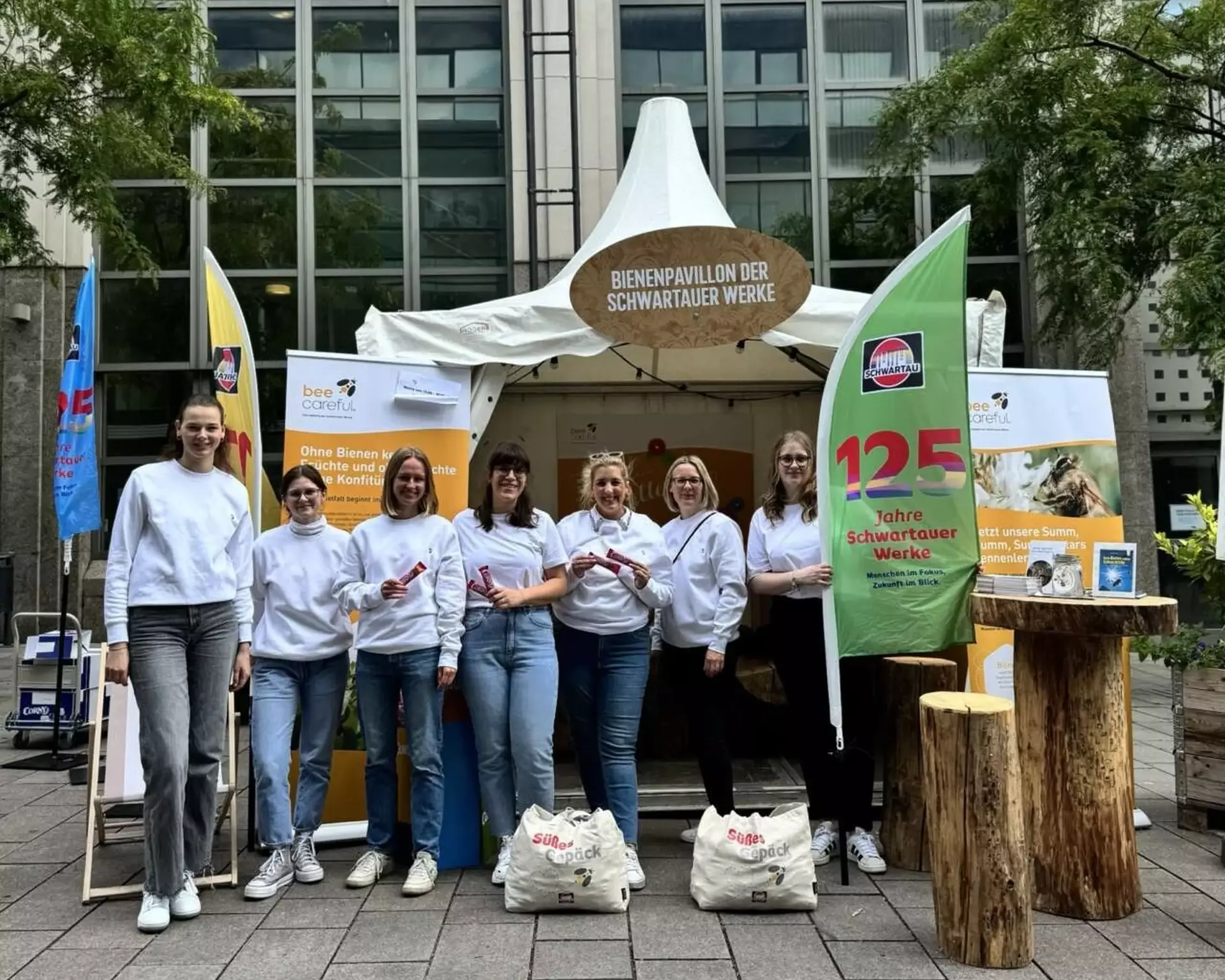 Man sieht eine Gruppe aus sieben jungen Frauen in einheitlichen weißen Sweatshirts vor einem großen Zelt stehen. Sie tragen Flaggen mit dem Slogan "125 Jahre Schwartauer Werke". Am Zelt ist auf einem großen Schild "Bienenpavillon" zu lesen, man sieht im Hintergrund Infotafeln und Holzmöbel.