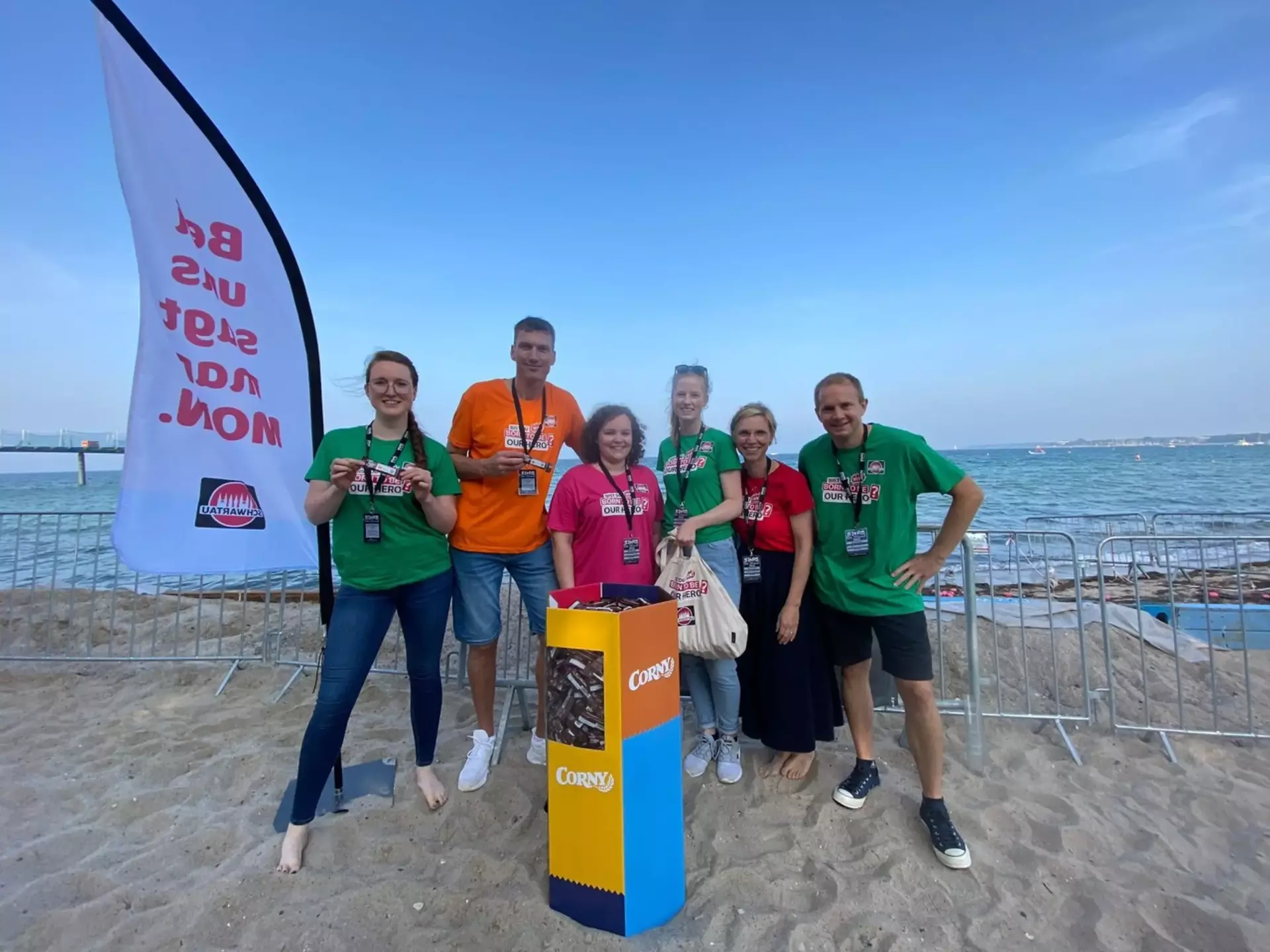 Man sieht zwei Männer und vier Frauen in bunten T-Shirts am Strand stehen. Vor ihnen steht eine bunte Schütte voller Corny-Riegel. Auf einer großen Flagge nehmen der Gruppe steht der Slogan "Bei uns sagt man Moin".