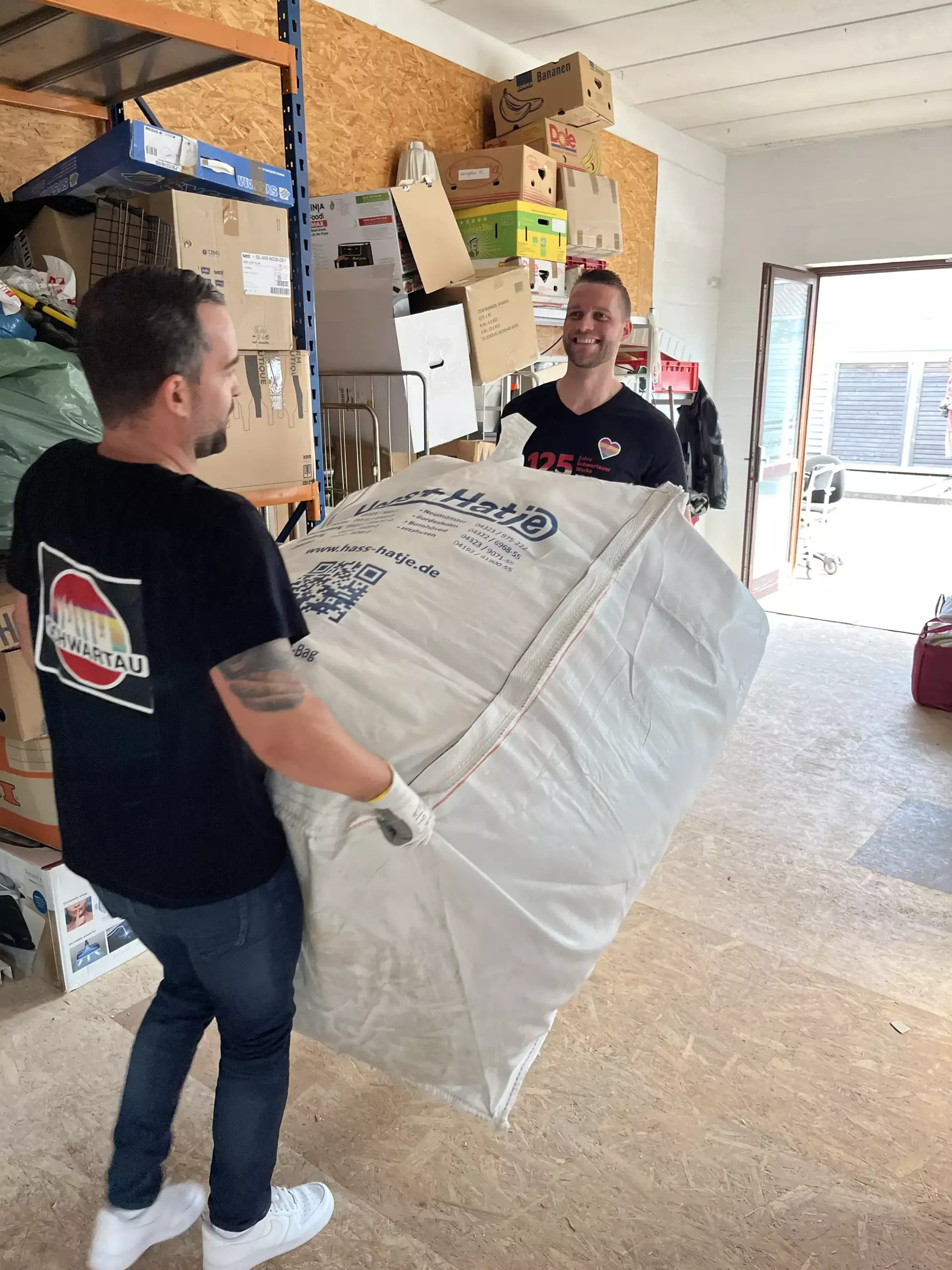 Dieses Foto zeigt zwei Schwartau Kollegen, die ein weißes Bigbag in einer Halle tragen und dabei in lächeln.