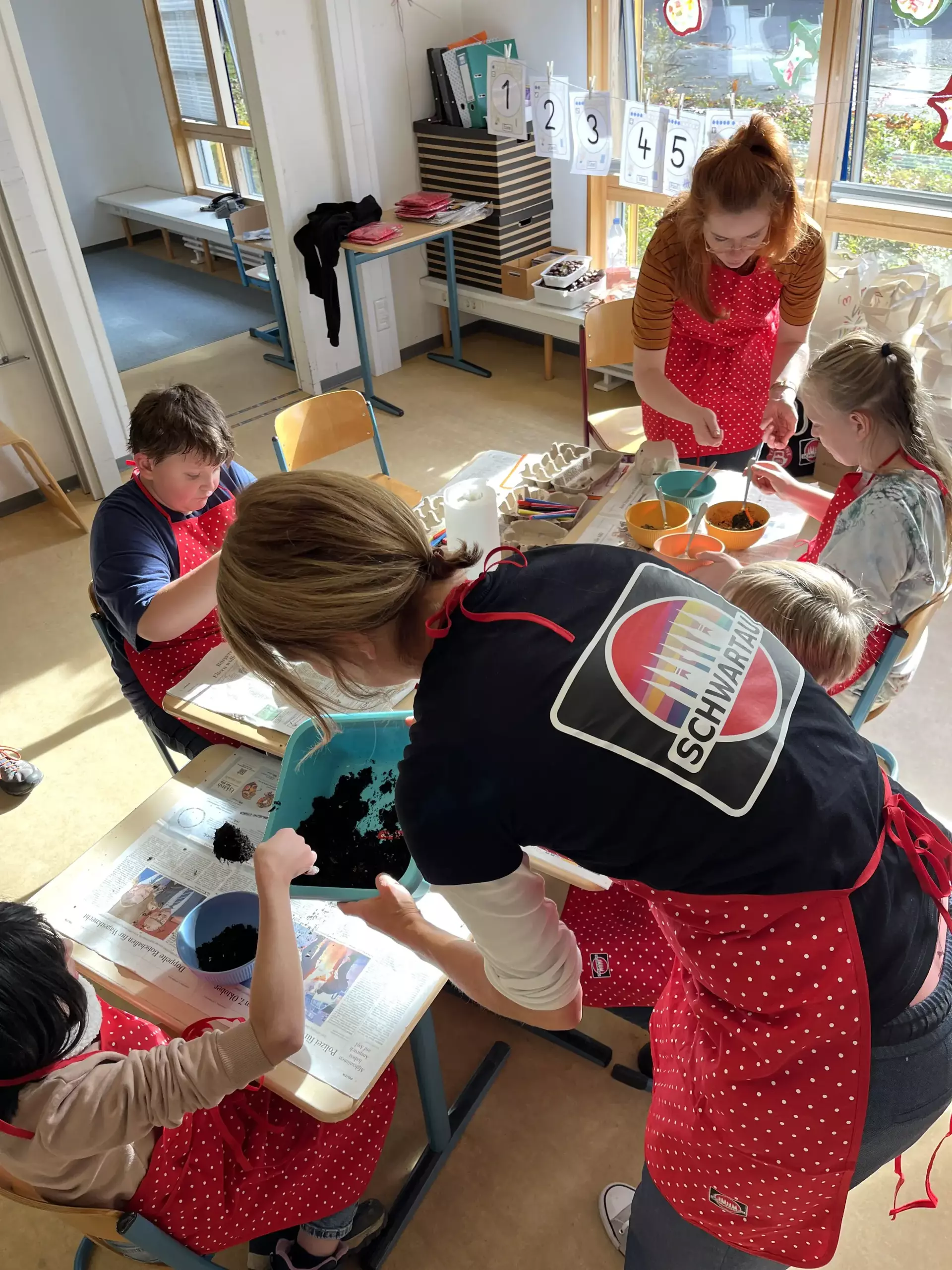 Dieses Bild zeigt eine Gruppe an Leuten, die Seedbombs basteln. Alle Personen tragen rote Schürzen mit Schwartau-Logo.