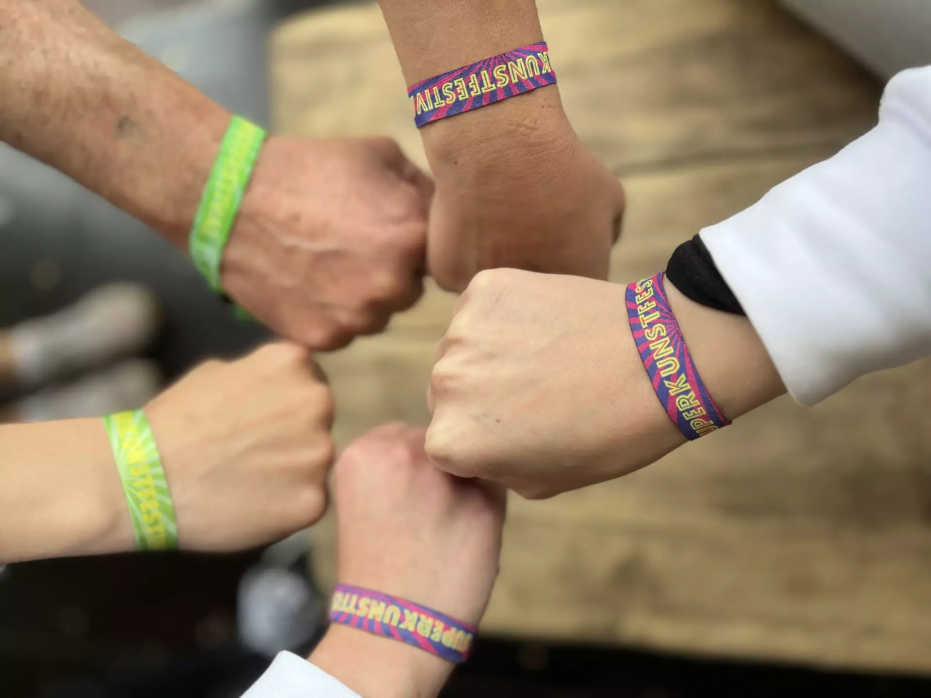 Man sieht vier Hände zur Faust geballt, die sich in der Mitte des Bildes treffen. Alle tragen am Handgelenk das Festivalbändchen des Superkunstfestivals