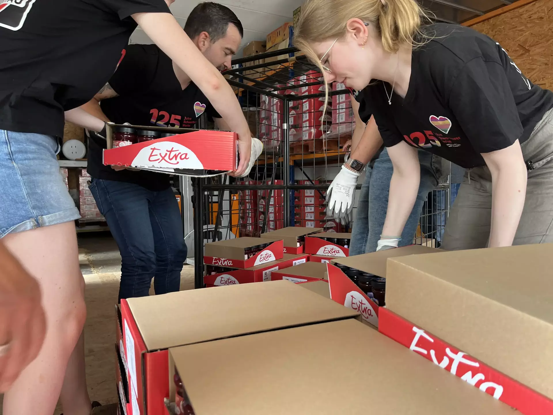 Das Bild zeigt vier Schwartau Kolleg*innen, die Konfitüren Trays in eine Gitterbox sortieren. Alle Personen und die Gitterbox steht in einem Lagerraum.