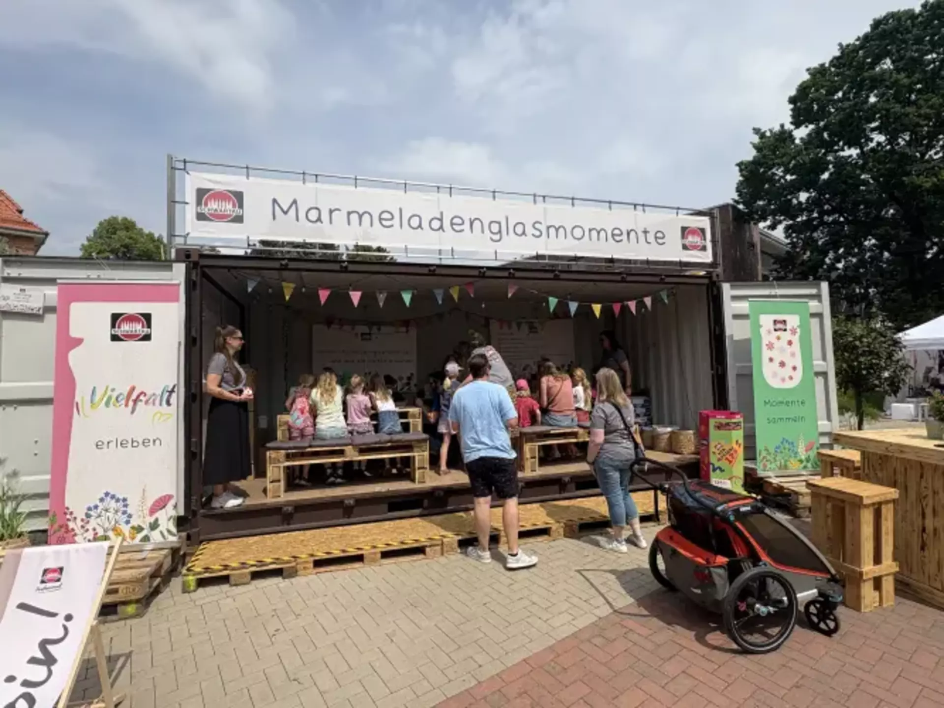Man sieht einen großen Stand in Form eines Schiffscontainers. Obendrüber ein großes Banner mit der Aufschrift Marmeladenglasmomente. Im geöffneten Container sieht man Kinder an Holzmöbeln sitzen und malen. An den Seiten stehen bunte Banner mit Marmeladengläsern.