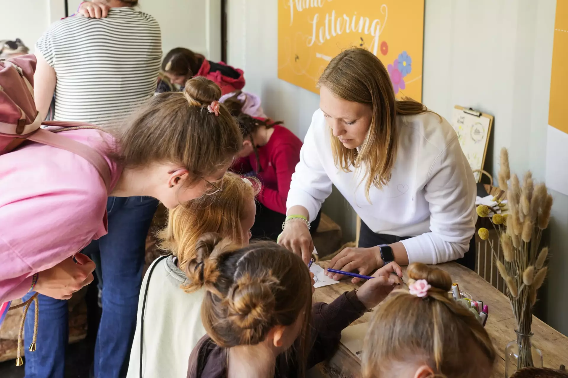 Im Vordergrund vier Mädchen von hinten an einem Tisch, davor eine junge Frau in weißen Sweatshirt, die Bastelmaterialien ausgibt.
