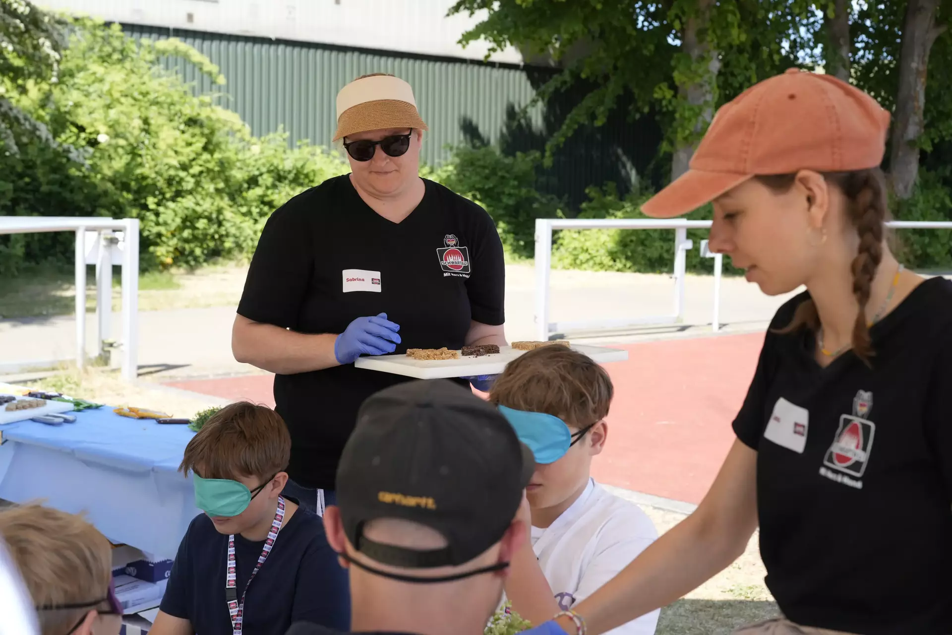 Zwei Mitarbeiterinnen in schwarzen Schwartau-Shirts stehen betreuend daneben und begleiten die blinde Verkostungsaktion. Im Vordergrund sind einige Kinder mit Augenklappen zu sehen.