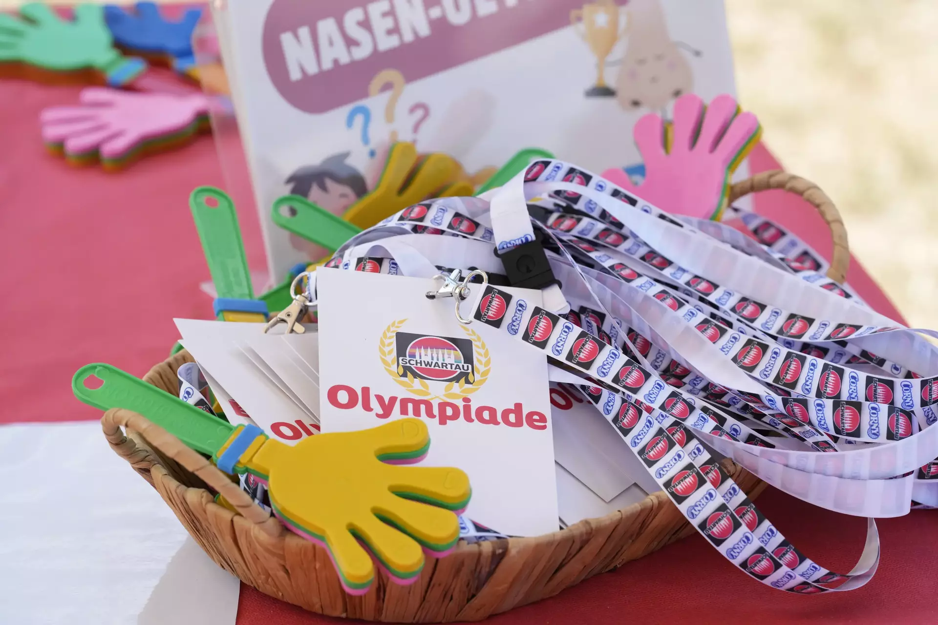 Auf dem Bild ist ein bunter und einladender Tischaufbau zu sehen, der zur Schwartau-Olympiade bei den Special Olympics gehört. Im Mittelpunkt steht ein geflochtener Korb, der mit Lanyards (Schlüsselbändern) mit dem Schwartau-Logo und großen Namenskarten mit der Aufschrift „Olympiade“ gefüllt ist. Die Lanyards sind weiß mit Schwartau-Logos in einer Reihe bedruckt. Außerdem befinden sich im Korb und drumherum bunte Klatschhände aus Schaumstoff und ein Aufsteller im Hintergrund mit der Aufschrift „NASEN-OLYMPIADE“, begleitet von lustigen Zeichnungen, z. B. einer Nase mit Pokal. Das Ganze wirkt spielerisch, freundlich und ist offensichtlich auf Kinder oder Jugendliche zugeschnitten – perfekt passend zur fröhlichen, inklusiven Atmosphäre der Special Olympics. Die Szene strahlt Spaß, Teamgeist und Engagement aus – mit viel Liebe zum Detail gestaltet.