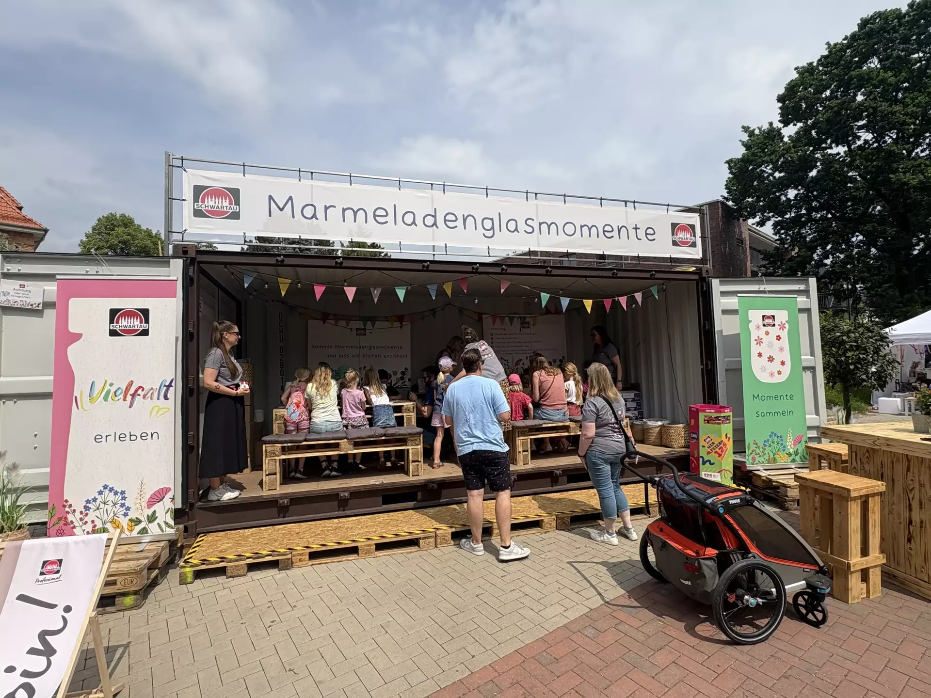 Man sieht einen großen Stand in Form eines Schiffscontainers. Obendrüber ein großes Banner mit der Aufschrift Marmeladenglasmomente. Im geöffneten Container sieht man Kinder an Holzmöbeln sitzen und malen. An den Seiten stehen bunte Banner mit Marmeladengläsern.