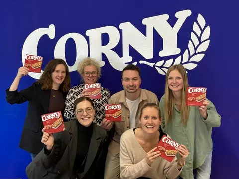 Mehrere Mitarbeitende des Corny-Teams stehen vor einer blauen Wand mit großem Corny-Logo und halten Verpackungen von Corny Cinnamon Roll in die Kamera.