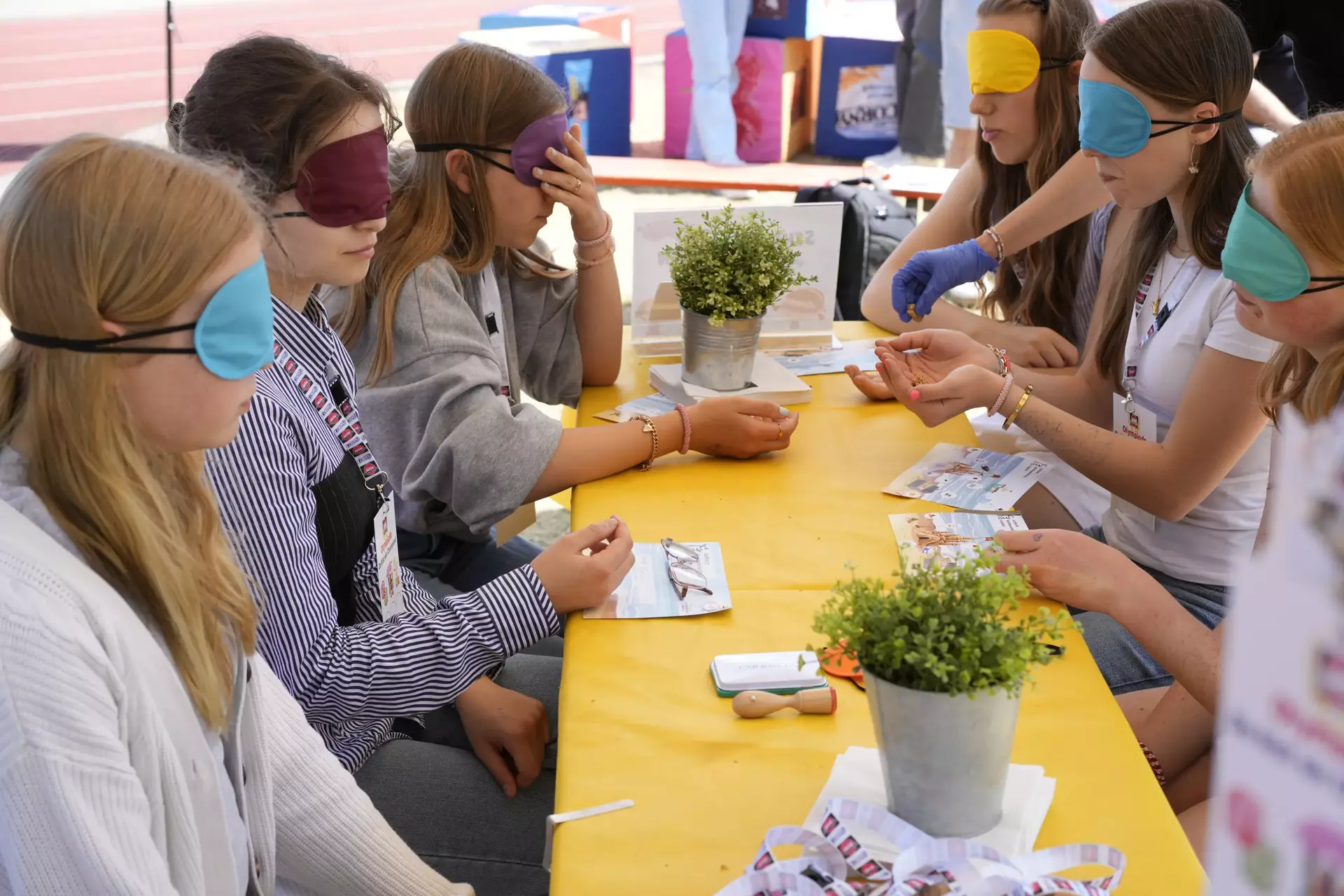 Auf dem Bild sieht man eine Gruppe junger Menschen, die an einem gelben Tisch sitzen – alle mit bunten Augenbinden. Es handelt sich um eine Station der Schwartau-Olympiade, vermutlich den „Schmeckathlon“, bei dem eine Blindverkostung stattfindet. Eine Person mit blauer Einweghandschuhen reicht gerade ein Produkt zum Probieren. Vor den Teilnehmenden liegen kleine Flyer, vermutlich zur Orientierung oder für Notizen. Die Szene zeigt konzentrierte Gesichter und eine ruhige, neugierige Atmosphäre. Durch die Augenbinden stehen Geschmack und Sinne im Mittelpunkt – ein kreativer Ansatz, um Barrieren spielerisch zu thematisieren und Erlebnisse zu schaffen, die verbinden. Im Vordergrund liegen wieder Schwartau-Lanyards, außerdem Topfpflanzen als freundliche Deko – alles liebevoll gestaltet. Das Bild strahlt Gemeinschaft, Teilhabe und Inklusion aus.