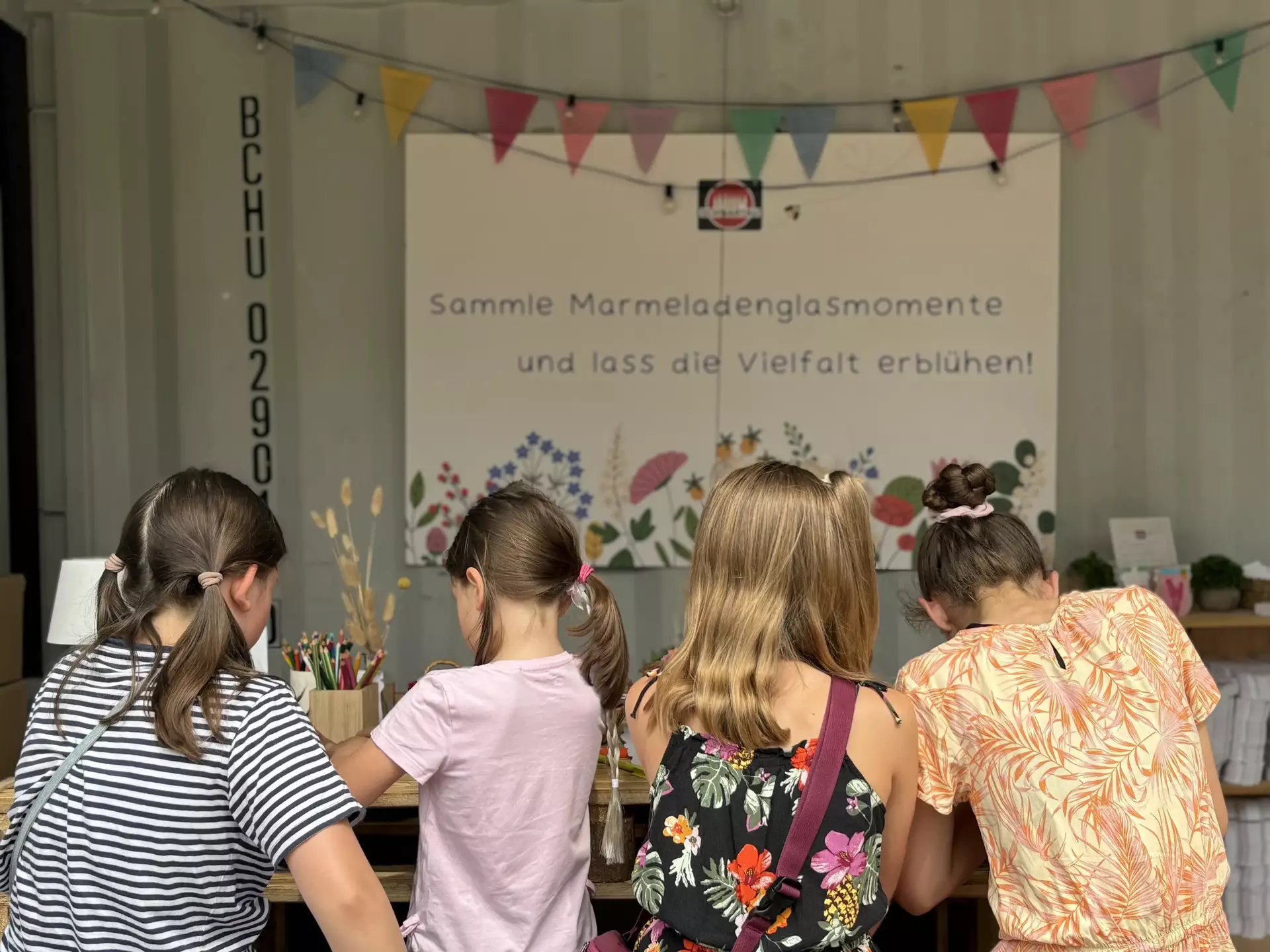 Man sieht vier Mädchen von hinten fotografiert an einem Holztisch sitzen und malen. Im Hintergrund hängt eine bunte Girlande und ein Schild mit der Aufschrift "Sammle Marmeladenglasmomente und lass die Vielfalt erblühen"