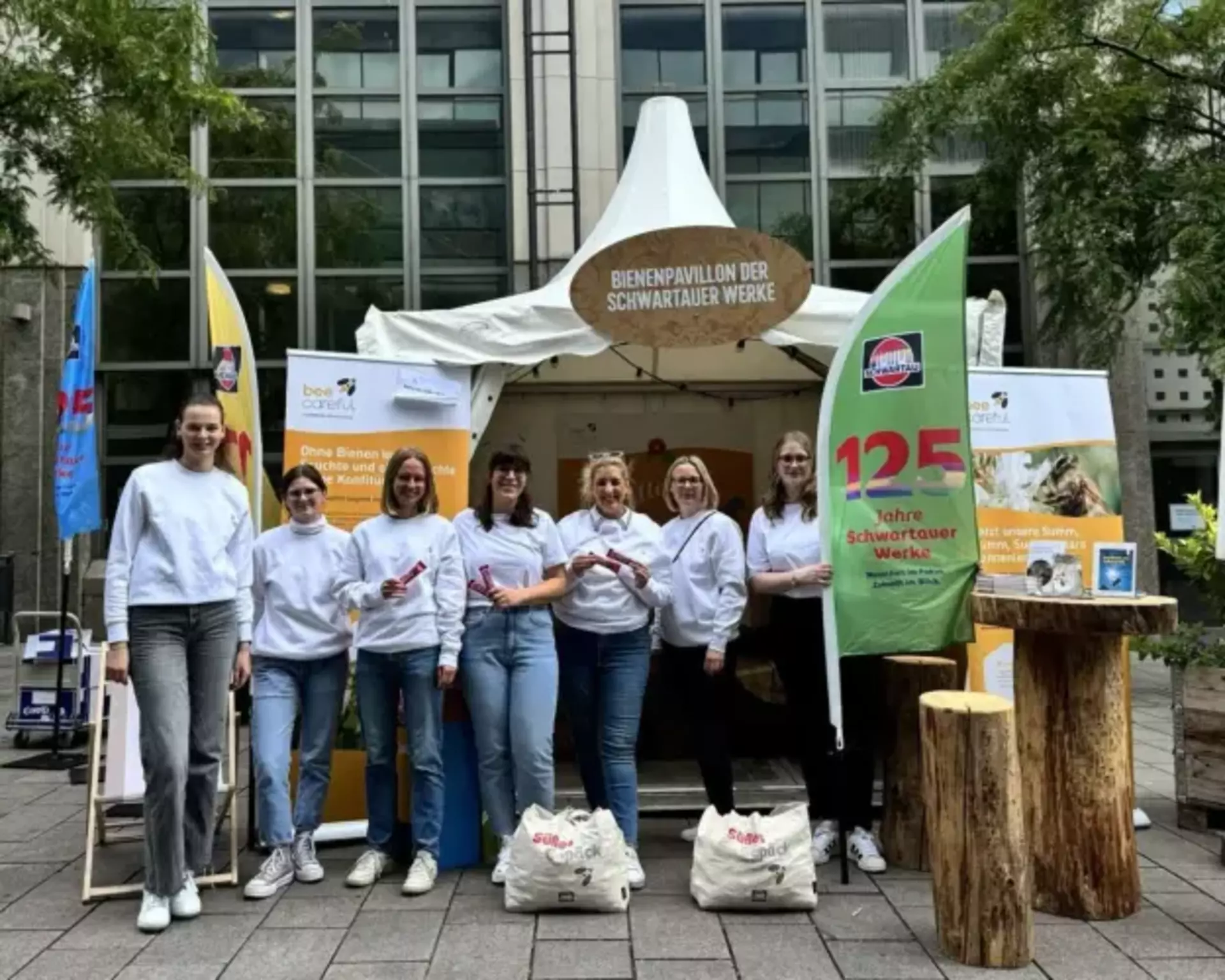 Man sieht eine Gruppe aus sieben jungen Frauen in einheitlichen weißen Sweatshirts vor einem großen Zelt stehen. Sie tragen Flaggen mit dem Slogan "125 Jahre Schwartauer Werke". Am Zelt ist auf einem großen Schild "Bienenpavillon" zu lesen, man sieht im Hintergrund Infotafeln und Holzmöbel.