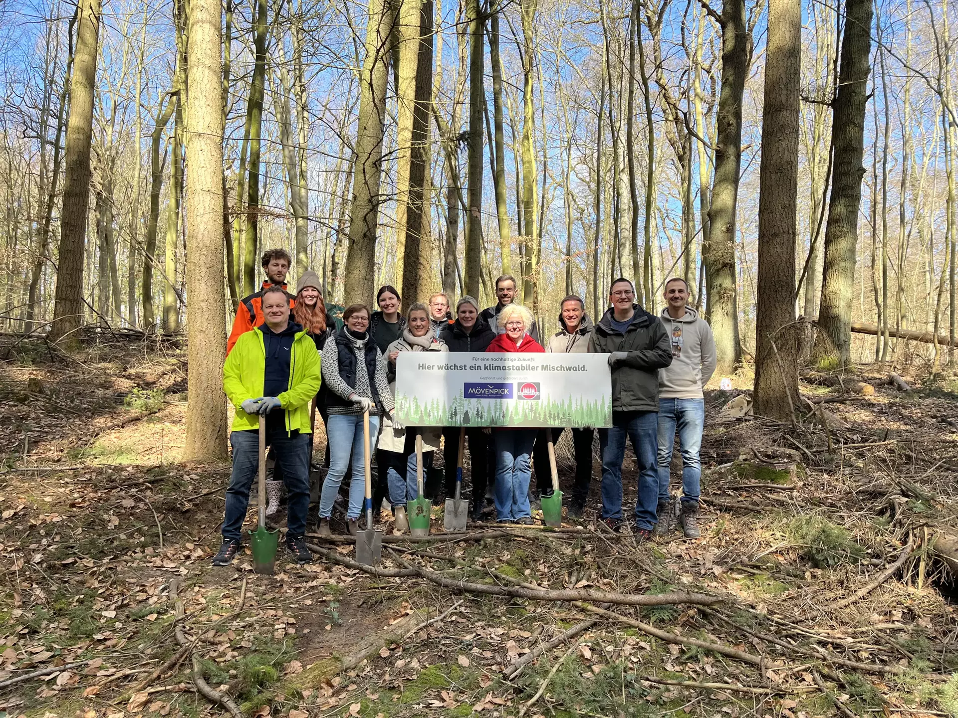 Zehn Kolleg*innen der Schwartauer Werke, drei von Mövenpick sowie die örtliche Försterei stehen im Kücknitzer Wald mit dem Schild "Hier wächst ein klimaneutraler Mischwald".
