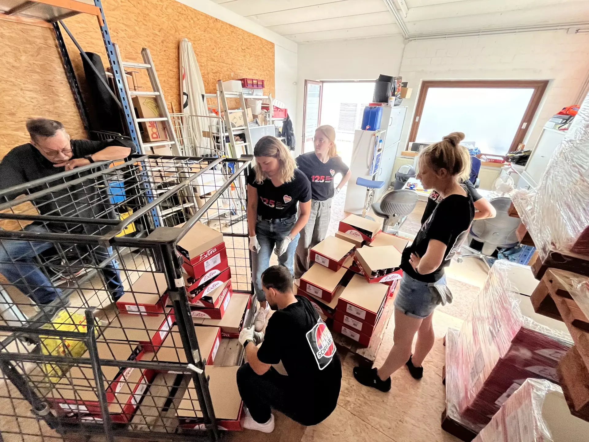 Das Bild zeigt fünf Schwartau Kolleg*innen, die Konfitüren Trays in eine Gitterbox sortieren. Auf der Rückseite der Gitterbox lehnt ein Mitarbeiter vom ASB. Alle Personen und die Gitterbox steht in einem Lagerraum.
