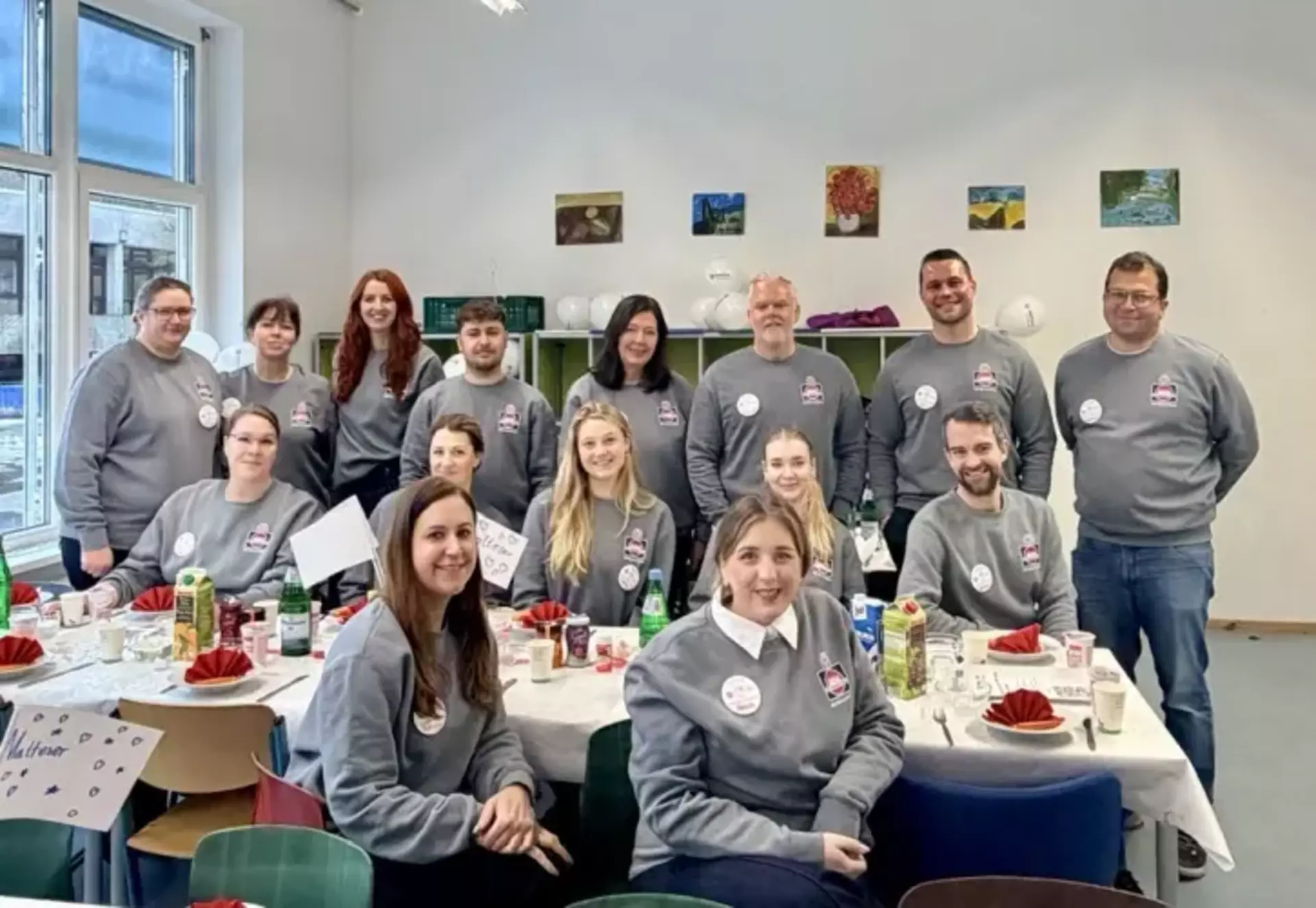 Gruppenfoto aller teilnehmenden Kolleg*innen, zu sehen sind 15 Personen unterschiedlichen Alters mit grauen Schwartau-T-Shirts, sitzend an einem großen gedeckten Frühstückstisch