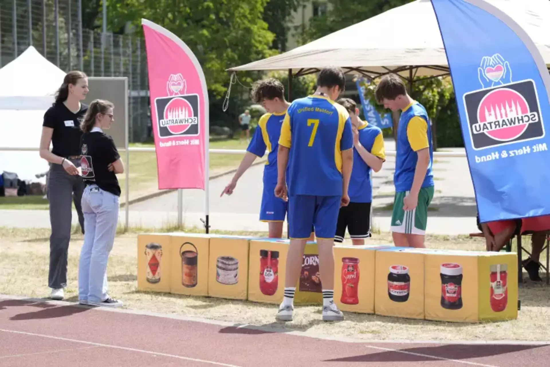Auf dem Bild ist eine sportliche und fröhliche Atmosphäre zu sehen. Eine Gruppe junger Menschen in blauen Trikots mit gelben Akzenten (Team „Unified Meldorf“) steht vor einer Spielstation der Schwartau-Olympiade. Die Teilnehmenden zeigen auf die abgebildeten Produkte – es scheint sich um die Station „Würfel-Spaß“ zu handeln, bei der Schwartau-Produkte in die richtige Reihenfolge gebracht werden sollen. Zwei Mitarbeiterinnen in schwarzen Schwartau-Shirts stehen betreuend daneben und begleiten die Aktion. Bunte Schwartau-Fahnen mit dem Slogan „Mit Herz & Hand“ rahmen das Bild ein und unterstreichen das Engagement des Unternehmens. Die Szene vermittelt Teamgeist, Aktivität und Spaß – kombiniert mit der klaren Botschaft: Inklusion leben, Gemeinschaft stärken.