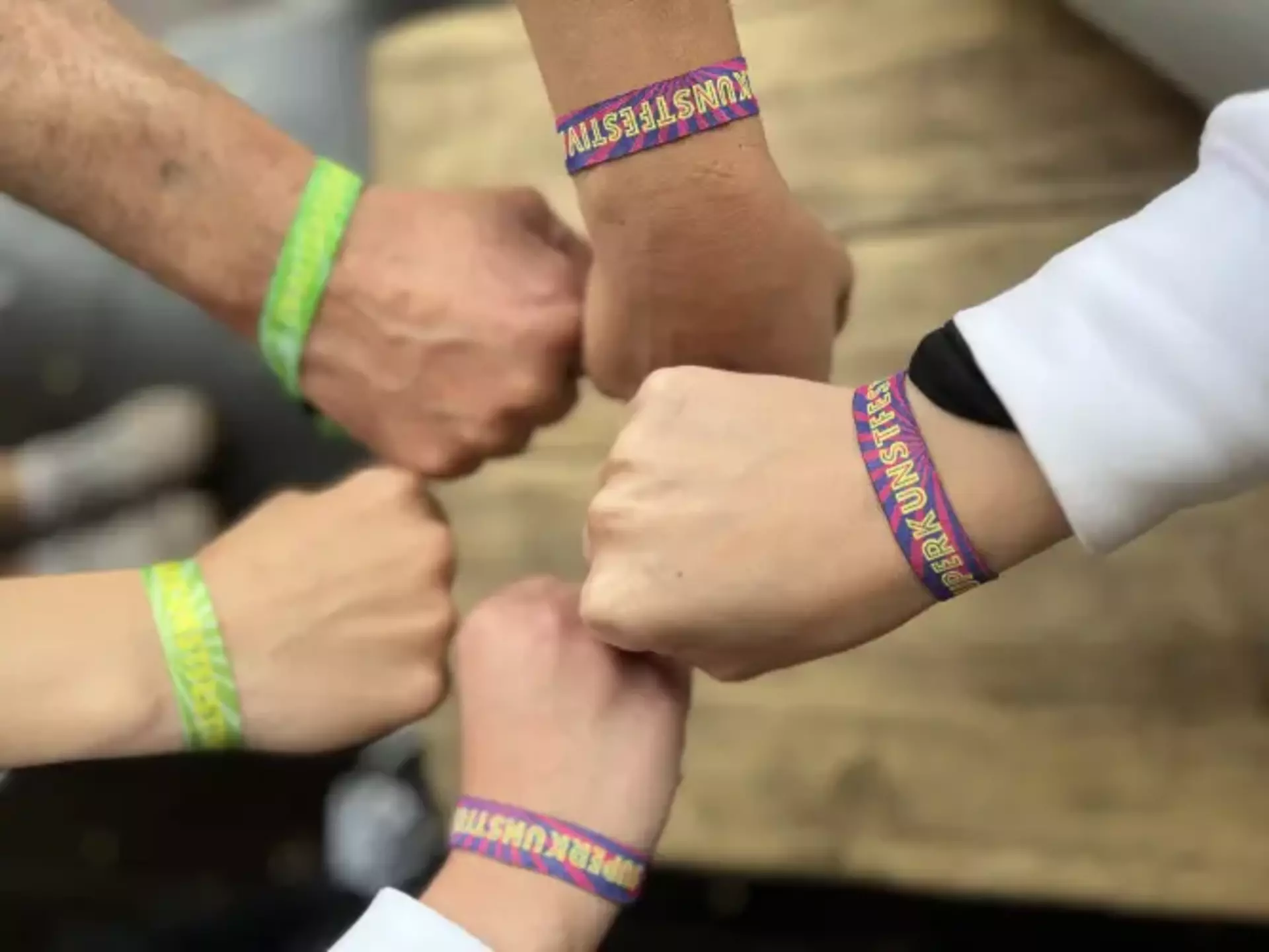 Man sieht vier Hände zur Faust geballt, die sich in der Mitte des Bildes treffen. Alle tragen am Handgelenk das Festivalbändchen des Superkunstfestivals