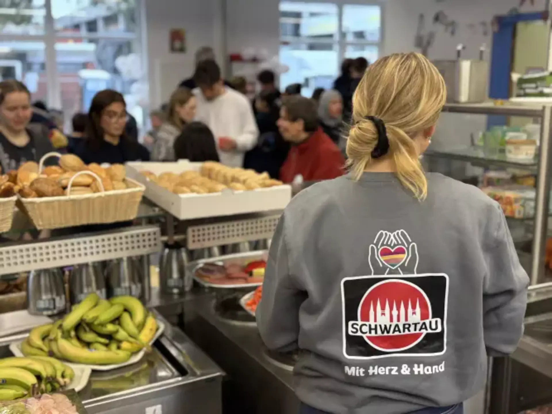 Man sieht den Rücken eine Kollegin mit dem großen Schwartau-Logo bedruckt am Ausgabe-Buffett, vorne Körbe mit Obst und Brötchen.