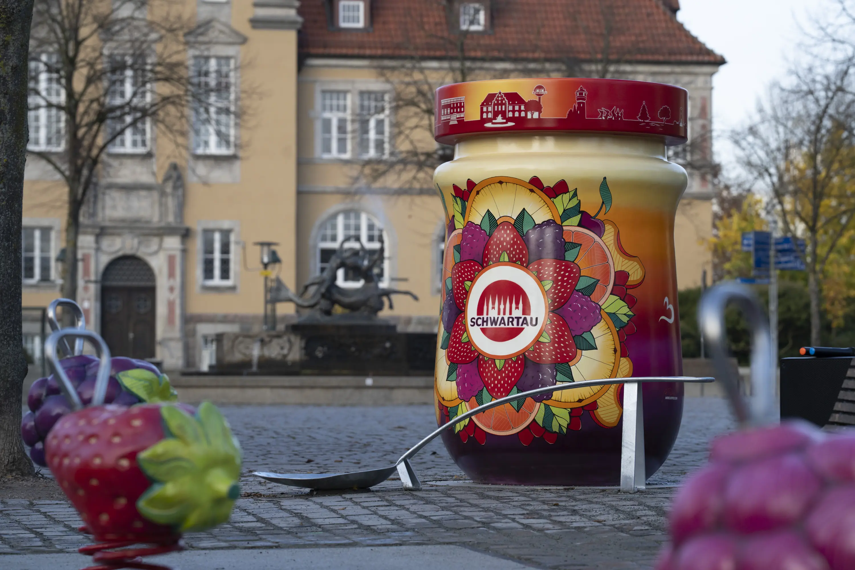 Auf dem Bild sieht an den Fotopoint "Marmeladenstadt" mit vielen bunten Farben, den Metalllöffel im Vordergrund des großen Marmeladenglases und zwei Federwippen in Form einer Erdbeere und Himbeere.