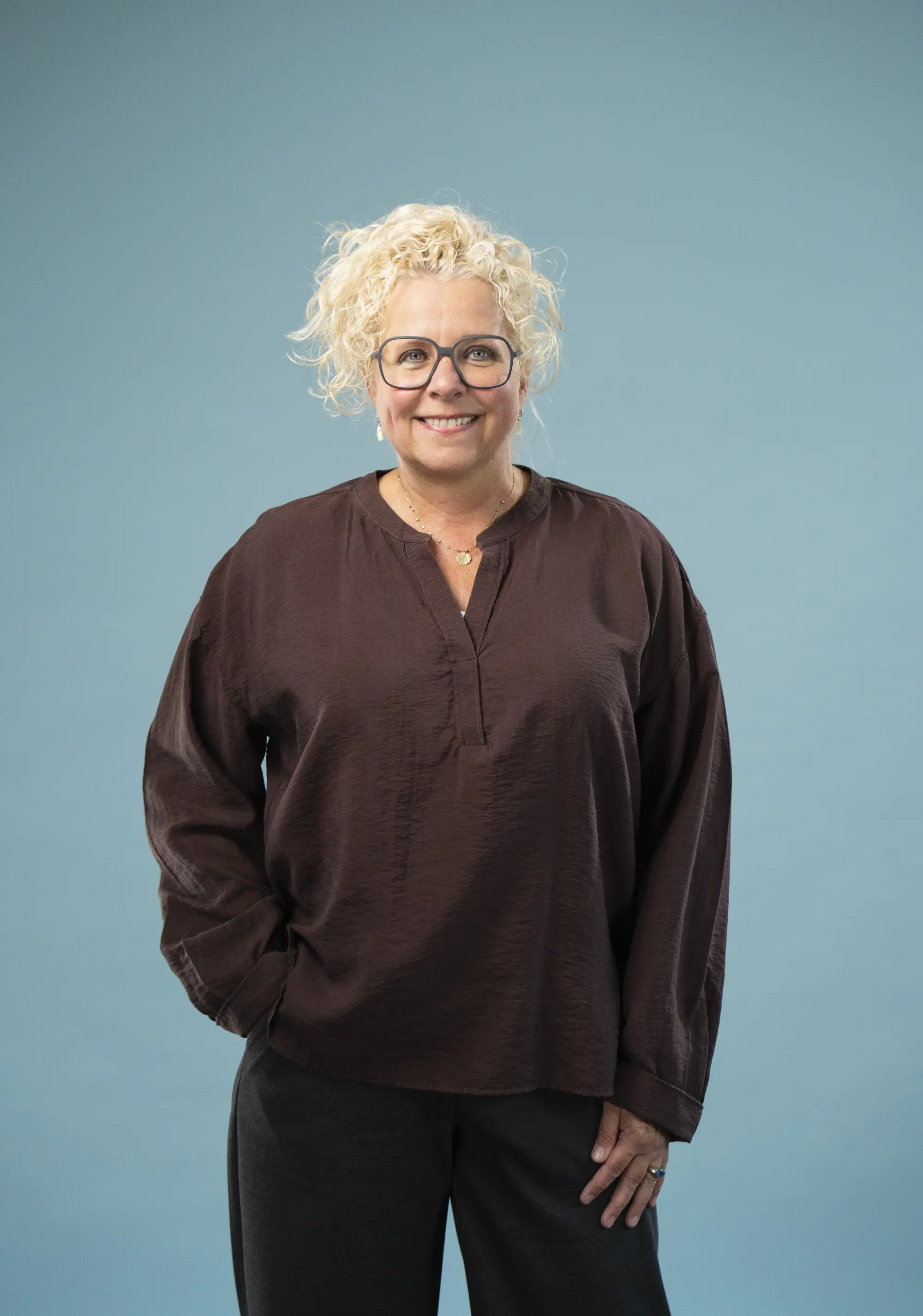 Eine Frau mit blonden Locken und einer Brille, in dunkler Hose und Bluse mit einem freundlichen Lächeln