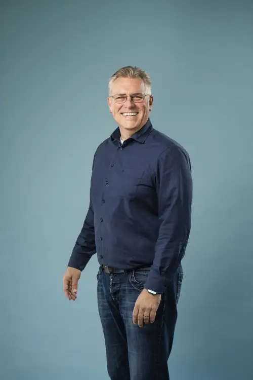 Ein Mann in dunkler Jeans und dunkelblauem Hemd vor einer Leinwand, graue kurze Haare und Brille mit freundlichem Lächeln