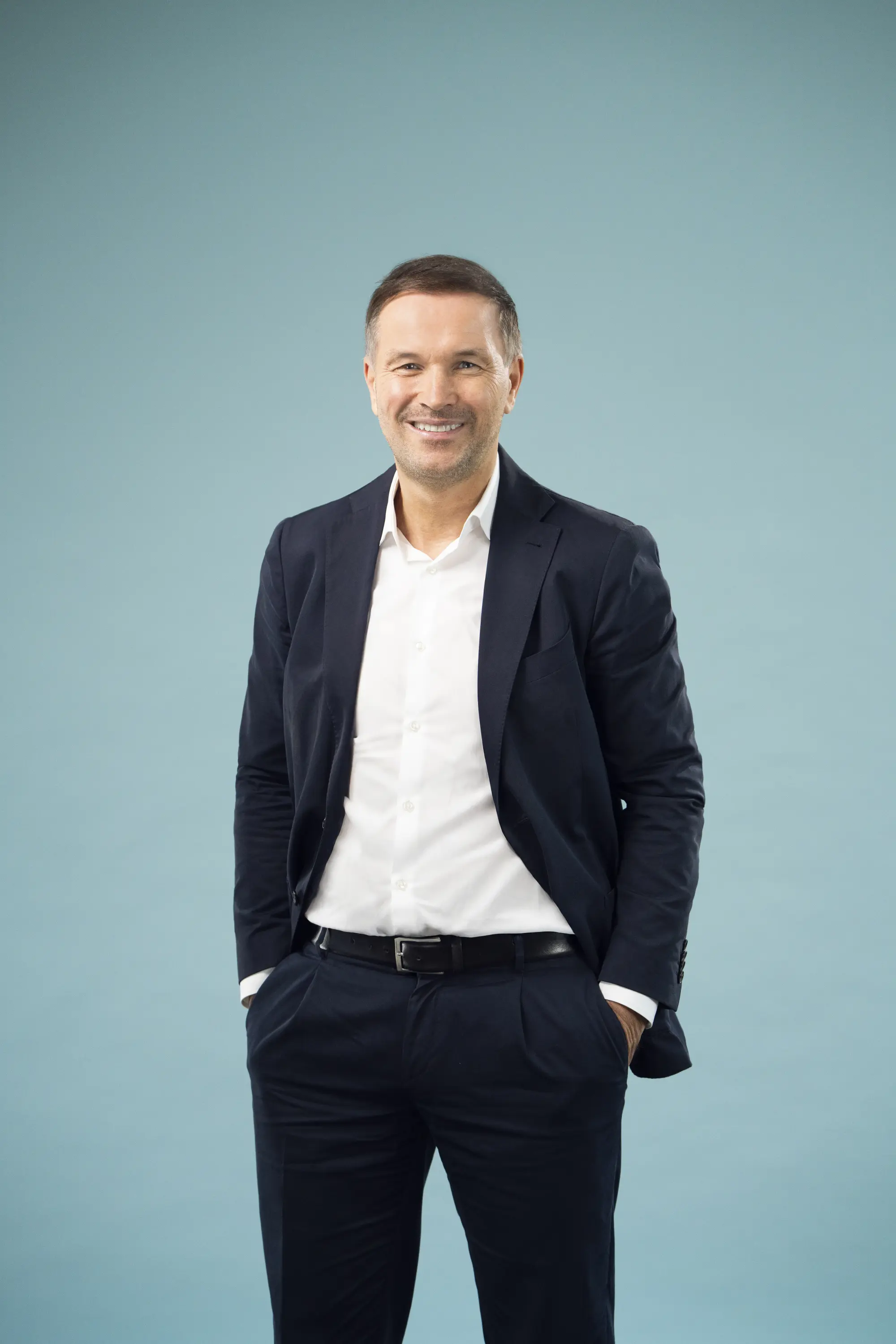 Mann in dunkler Hose und Sacko mit weißem Hemd vor einer Leinwand, freundliches Lächeln