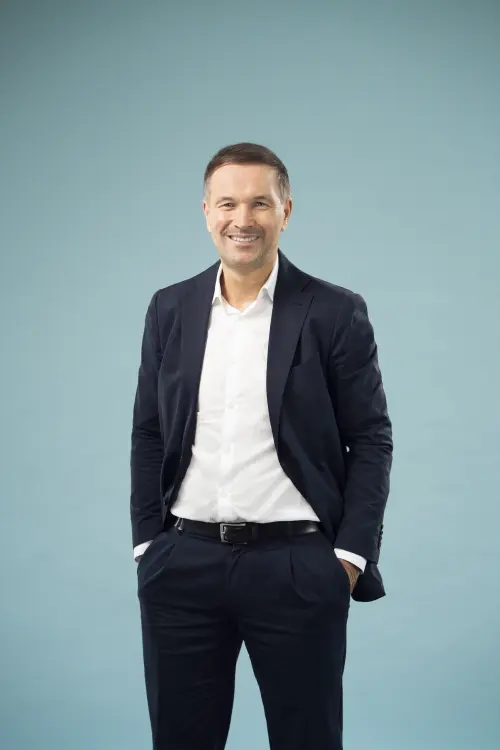 Mann in dunkler Hose und Sacko mit weißem Hemd vor einer Leinwand, freundliches Lächeln