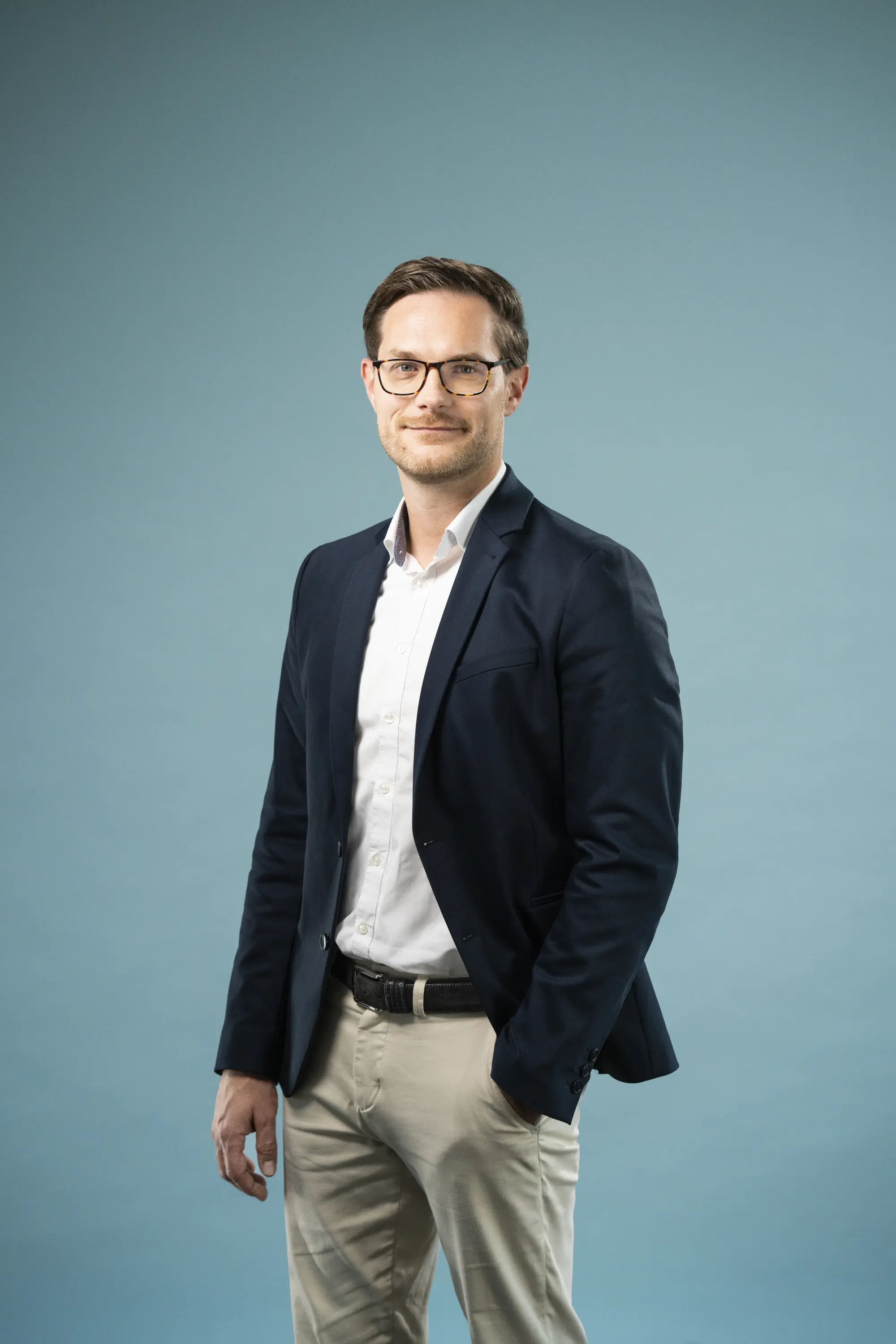 Schlanker Mann in Chino und Sacko mit Brille vor einer Leinwand