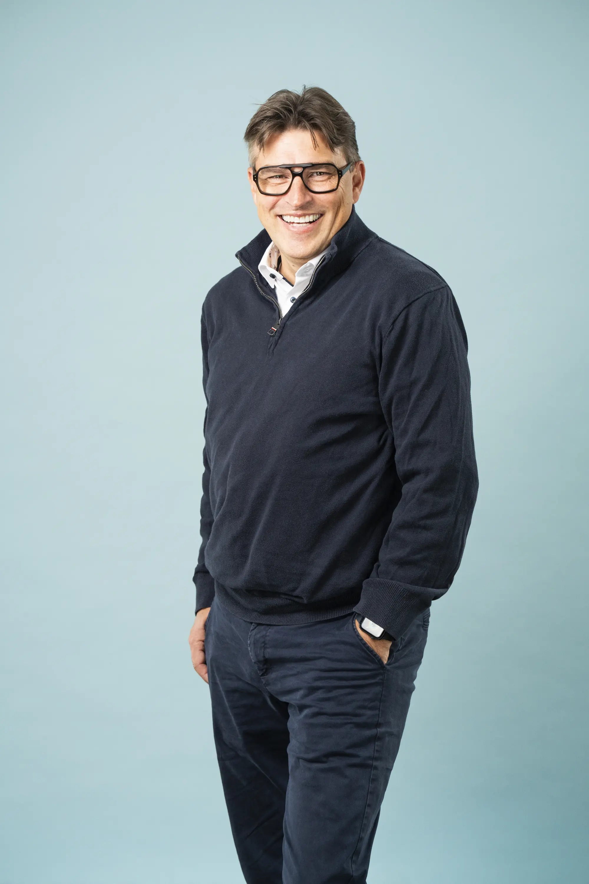 Schlanker Mann mit Brille, dunkle Hose & Pullover vor einer Leinwand, freundliches Lächeln