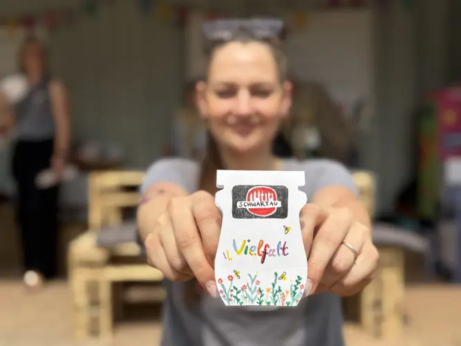 Man sieht unscharf eine junge Frau mit dunklen Haaren, sie hält eine bemalte Pappschachtel in Form eines Marmeladenglases in die Kamera. Auf dem Glas sind gemalte Blumen und das Wort Vielfalt und das Schwartau Logo zu sehen. Im Hintergrund des Bildes sieht man einen Stand mit Holzmöbeln.