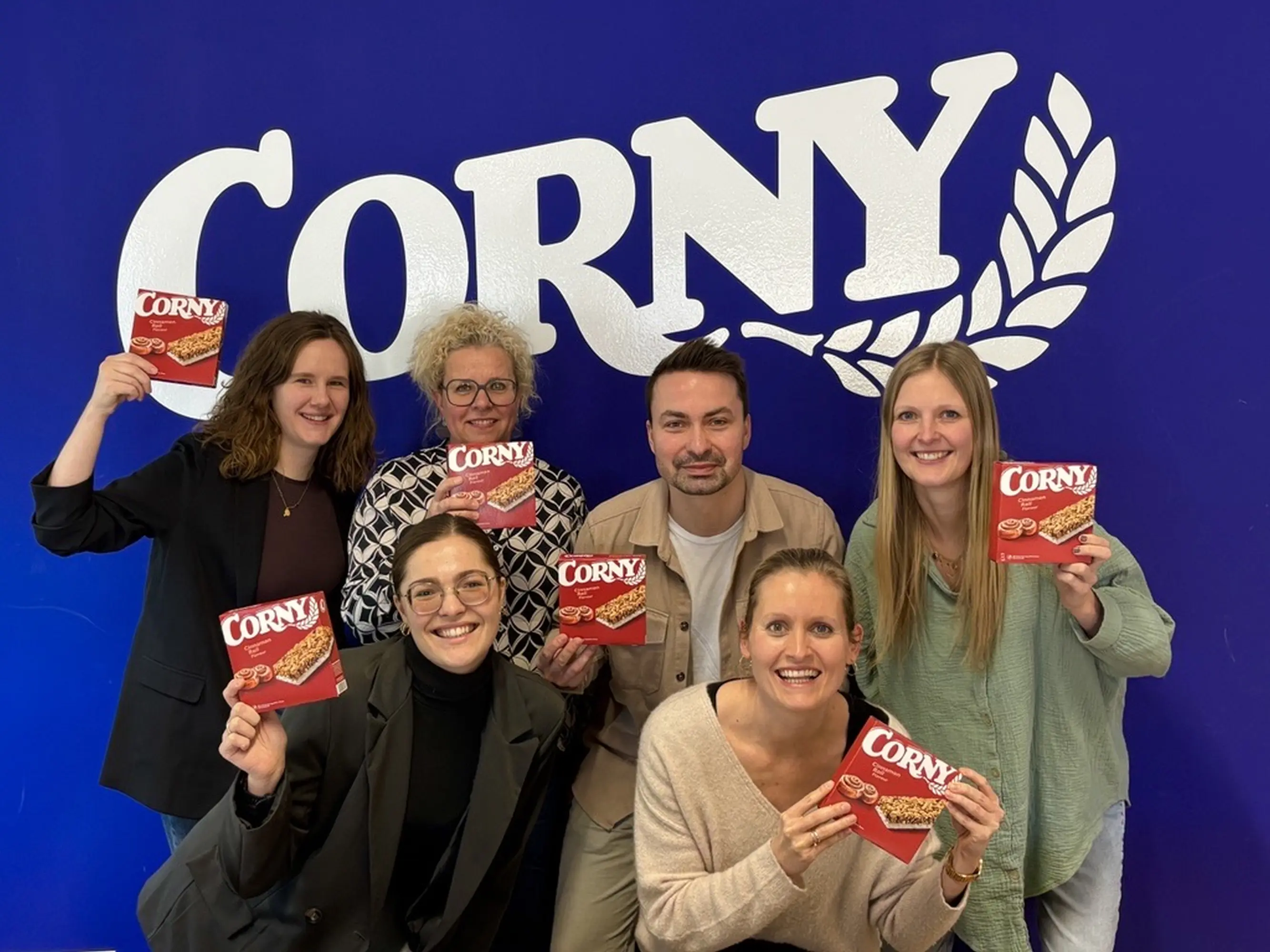 Mehrere Mitarbeitende des Corny-Teams stehen vor einer blauen Wand mit großem Corny-Logo und halten Verpackungen von Corny Cinnamon Roll in die Kamera.