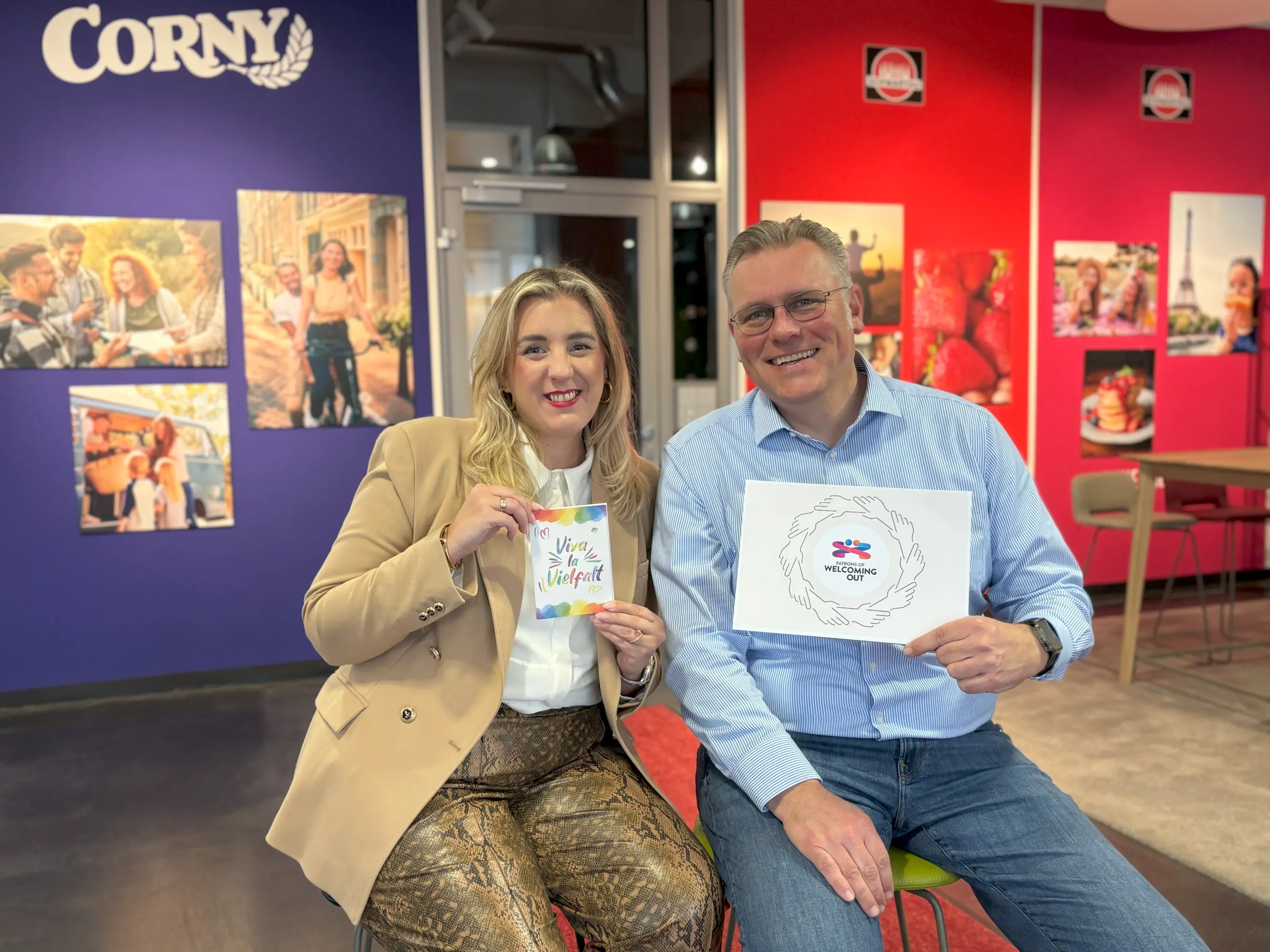 Die Teamleiterin Unternehmenskommunikation Vanessa Krabiell & der HR Director Florian Brombach sitzen nebeneinander im farbenfrohen Ein der Schwartauer Werke mit den Marken Corny & Schwartau im Hintergrund. Vanessa links hält eine Karte mit der Aufschrift „Viva la Vielfalt“, Florian rechts hält ein Blatt mit dem Logo der Initiative „Welcoming Out“. Im Hintergrund sind Wandbilder und das Corny-Logo sowie das Schwartau-Logo zu sehen.