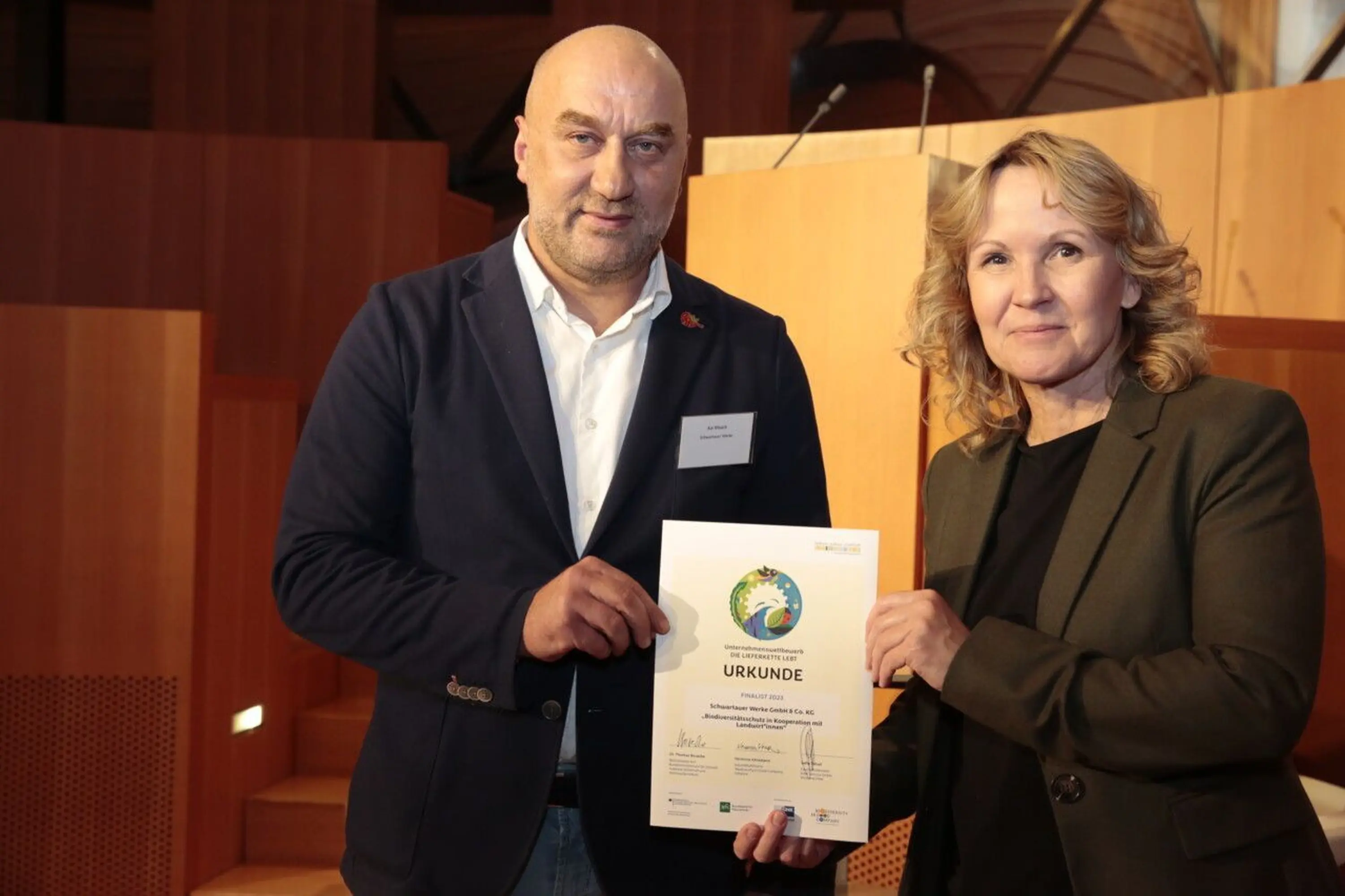 Das Bild zeigt zwei Personen, die schick angezogen sind und eine Urkunde überreichen. Auf der linken Seite steht unser Kollege Kai Maack, welcher von der Umweltministerin Steffi Lemke die Urkunde für "Die Lieferkette lebt" überreicht bekommt.
